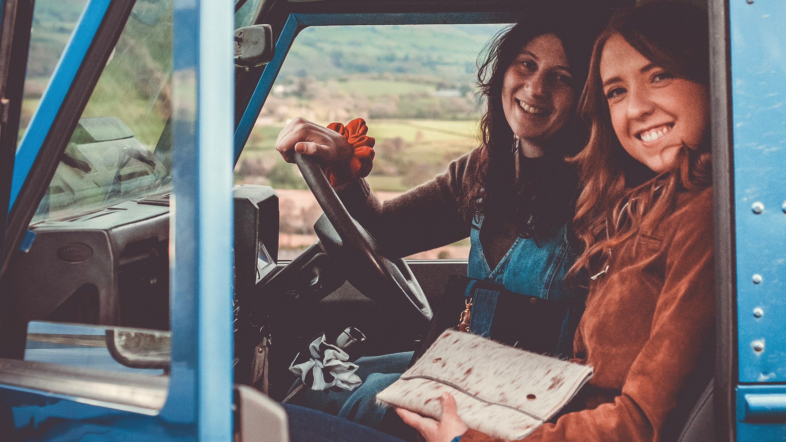 Two women sitting inside a vintage blue truck with a scenic background holding a handmade leather cowhide bag